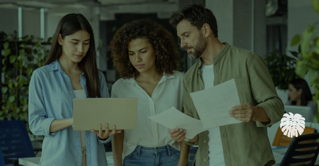 Three coworkers in an office collaborate on reviewing documents and a laptop, with a focused central team member, representing the process of reviewing and managing workplace communications.