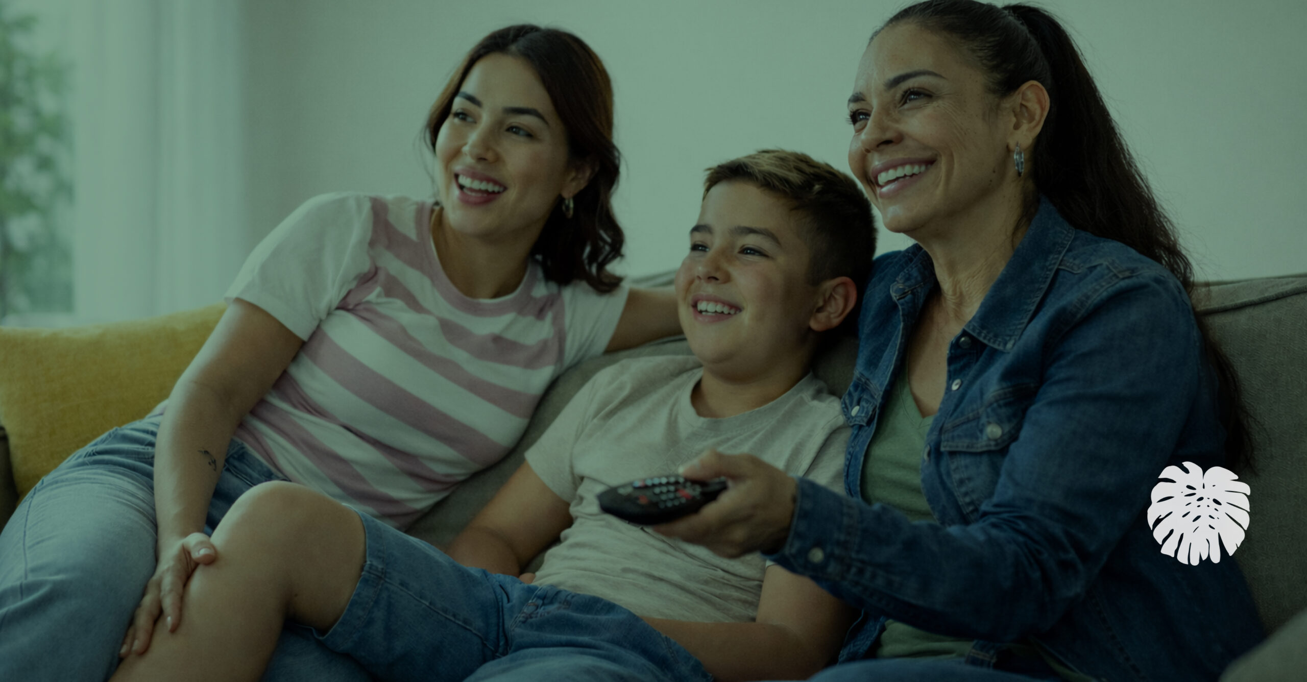 Hispanic mother, young woman, and boy sitting together on a couch in a bright living room, smiling and watching television, with the mother holding a remote control.